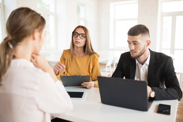Business man with laptop thoughtfully looking aside while business woman in eyeglasses with folder in hand talking with applicant about work. Young employers spending job interview in office