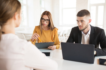 Young business man with laptop and business woman in eyeglasses thoughtfully looking at applicant. Young employers spending job interview in office