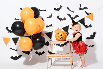 Fototapeta premium Baby girl in halloween costume with balloons and pumpkin bucket on white background