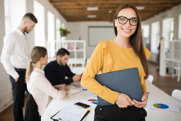 Young smiling business woman in eyeglasses happily looking in camera while holding folder in hands in office with colleagues on background