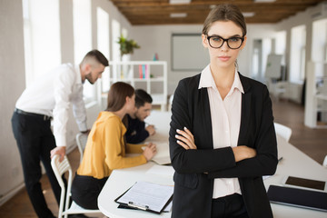 Young serious business woman in eyeglasses and shirt thoughtfully looking in camera while working in office with colleagues on background