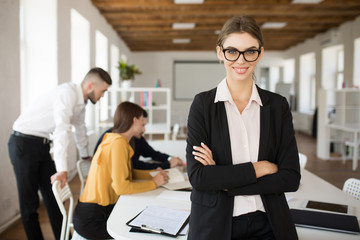 Young smiling business woman in eyeglasses and shirt happily looking in camera with folded hands while spending time in office with colleagues on background