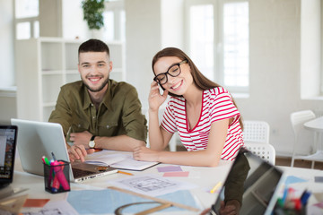 Pretty smiling girl in striped t-shirt and eyeglasses happily closing eyes while working with colleague. Group of cool guys with laptop spending time in modern office