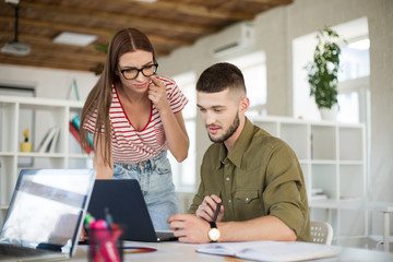 Young thoughtful man in shirt and woman in striped T-shirt and eyeglasses dreamily working together with laptop. Creative business people spending time at work in modern office