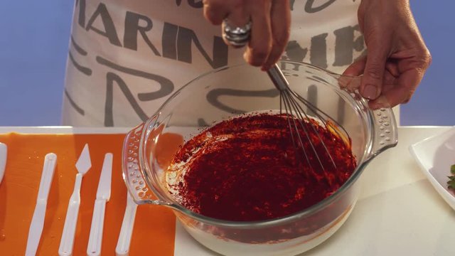 Slow motion demonstration of hand whisking of dark red substance in glass bowl with wide edges put on white table with set of different plastic knives and other cooking stuff.
