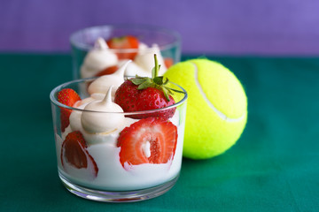 Wimbledon inspired whipped cream, meringues and fresh strawberries in a glass bowl on a green and violet background with a tennis ball