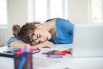 Young tired woman with eyeglasses on head sleeping on desk while working in modern office