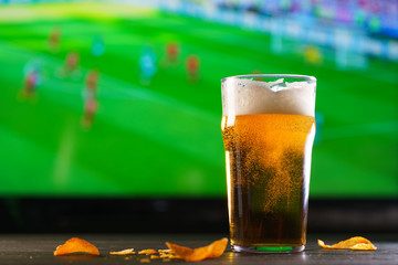 A glass of beer on a dark wooden table with crushed potato chips. Football on a background, high resolution