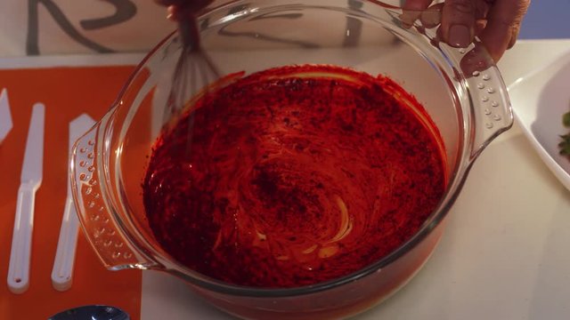 Hand whisking of red and white substance in glass pan with wide edges is turning it into orange color, put on table with set of white plastic knives and metallic spoons plus other cooking stuff.