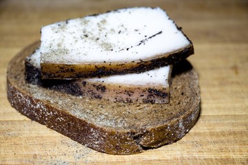 On a piece of black bread, there are two pieces of fat. The young pig's fat is sprinkled with black pepper spices. All this lies on a wooden surface.