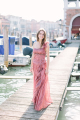 Beautiful young woman in rose dress standing on the background of Venice canal, buildings and gondola. Italy, Europe.
