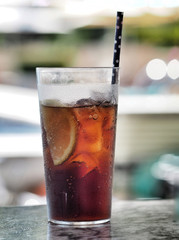 Cola with ice cube, slice of lemon and straw in glass.