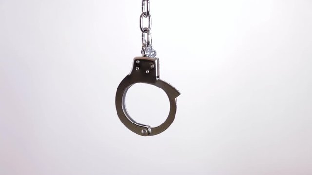 A pair of shiny metal handcuffs over a white background, suspended in the air. Symbolic shot: crime, punishment, menace, fetish, sexuality, pleasure.
