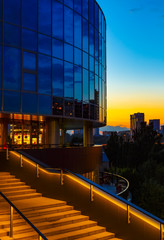 Naklejka na ściany i meble sunset on the background of round glass office building with a beautifully illuminated river and blue sky staircase,