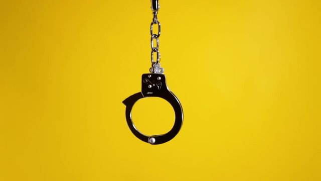 A pair of shiny metal handcuffs over a yellow background, suspended in the air. Symbolic shot: crime, punishment, menace, fetish, sexuality, pleasure.
