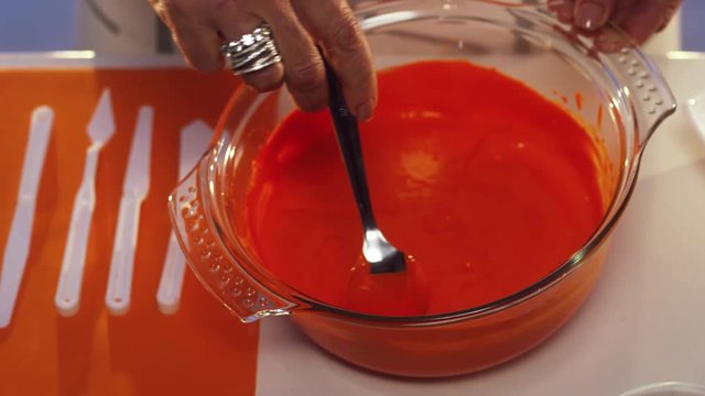 Woman's hand wearing rings stirs orange substance in big glass bowl with wide edges, put on white table with set of different sized knives and other cooking stuff.