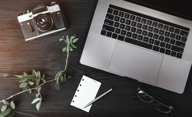 Close-up image of laptop, photo camera, eyeglasses, rose branch and white sheet on black wooden table. Top view