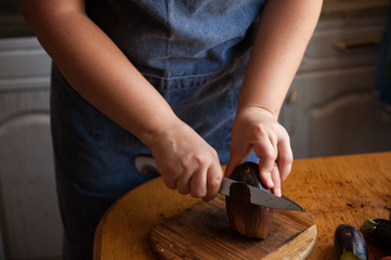 Woman cook in apron and cap cooks in kitchen, cuts eggplants, peppers, onions, garlic on cutting board with large knife. Stir with ladle in saucepan seasoning for canning. Hands close up