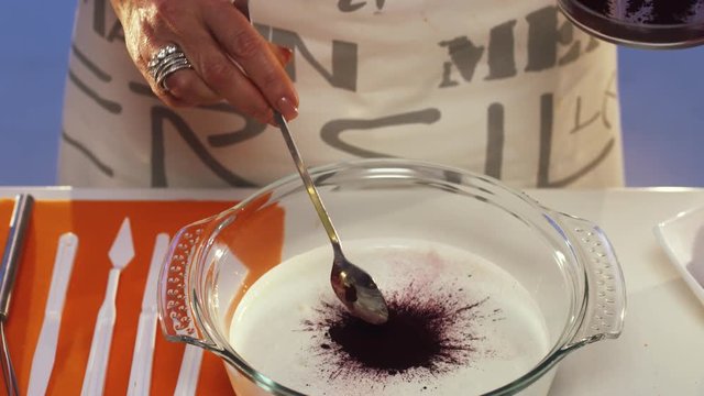 Slow motion of dark purple flour being sprinkled from small plate onto sour cream in big glass pan on white table beside dish with strawberries and set of spoons with forks and other cooking stuff.