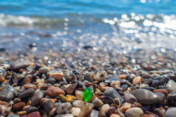 Green polished glass pebble on sea beach / Small green polished glass stone among pebbles on sea beach on background of splashing sea in sunny summer day