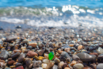 Green polished glass pebble on sea beach /	Small green polished glass stone among pebbles on sea beach on sunny summer day