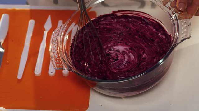 Slow motion view of hand whisking of violet and white substance in big round shaped glass bowl with wide edges put on white table with set of defferent plastic knives and other cooking stuff.