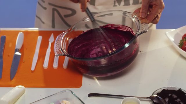 Showing of hand whisking of violet substance in big round shaped glass bowl with wide edges put on white table with set of defferent plastic knives, orange napkin and other cooking stuff.