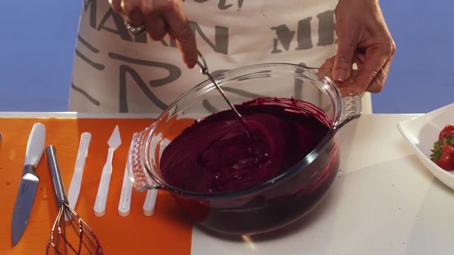 Process of hand whisking of violet substance in big round shaped glass pan with wide edges put on white table with set of defferent sized knives, orange napkin, additional whisk and other cook stuff.