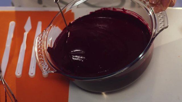 Woman in white ang grey apron is hand whisking violet substance in big round shaped glass pan with wide edges put on white table with set of defferent sized knives, additional whisk and orange napkin.