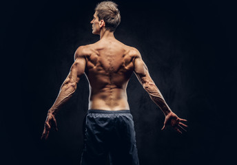 Back view of shirtless man with stylish hair and muscular ectomorph posing on the dark textured background.