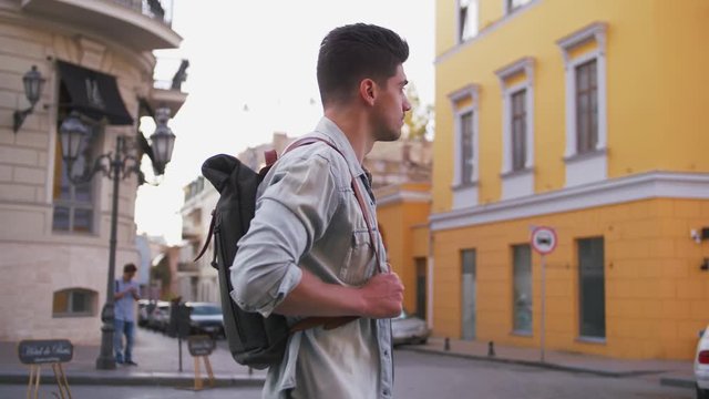 Attractive young handsome man tourist with backpack walking in city center and looking around, slow motion