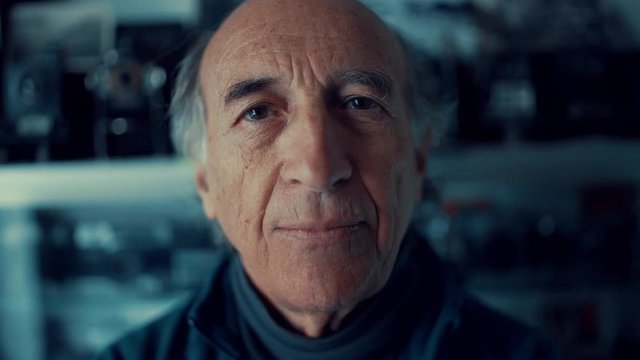 An old Caucasian man smiles at the camera in his shop of vintage cameras