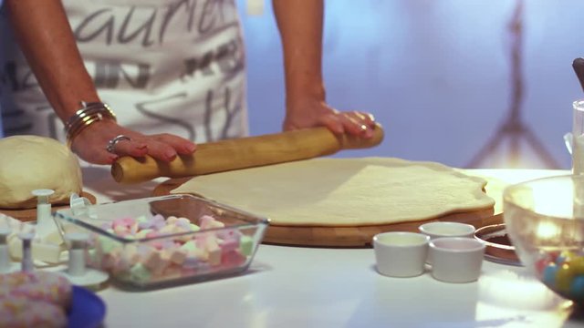 Woman in her late 40s dressed in colorful apron is rolling out dough on large round shaped wooden board with rolling pin on white kitchen table with defferent cooking stuff and blue background.