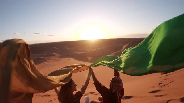 Women hold up cloth during desert sunset, slow motion