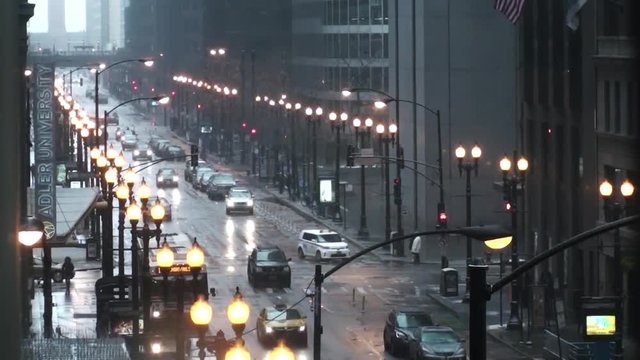 A rainy downtown Chicago intersection with medium traffic with mostly cars, buses and pedestrians. You see the headlights reflecting on the wet road surface and the street lights are lit.