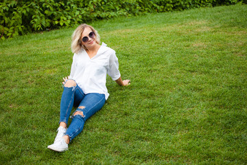 Fototapeta premium beautiful young smiling girl in sunglasses, white blouse and blue jeans sits on the grass