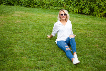 beautiful young smiling girl in sunglasses, white blouse and blue jeans sits on the grass