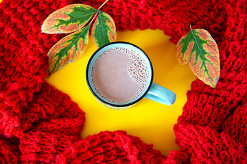Cocoa with milk in a blue ceramic mug, on a yellow background, surrounded by a red knitted scarf