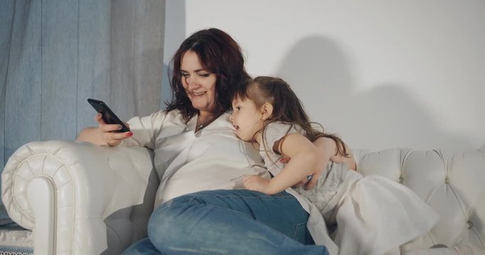 Pregnant mother and her little daughter are watching pictures on a smartphone.