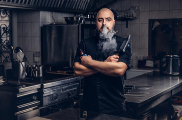 Bearded chef in black uniform smoking e-cigarette while standing in the kitchen.