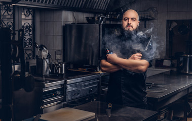 Bearded chef in black uniform smoking e-cigarette while standing in the kitchen.