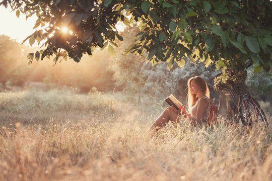 Beautiful young girl student sitting in a park under a tree reading a book.