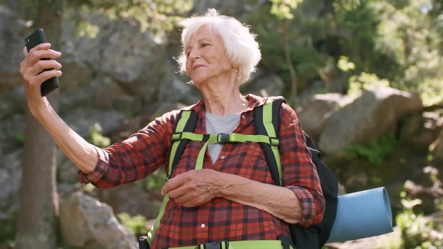 Beautiful senior woman with backpack smiling and taking selfie with smartphone while hiking in mountains