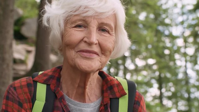 Portrait of beautiful senior woman looking at camera and smiling while standing with backpack in forest