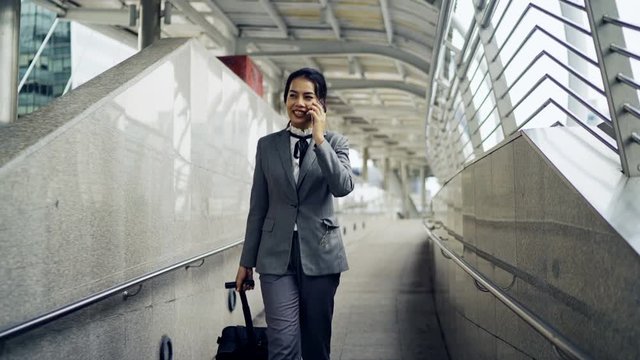 Asian business woman talking the mobile phone while dragging a wheeled luggage at the airport ramp for business trip