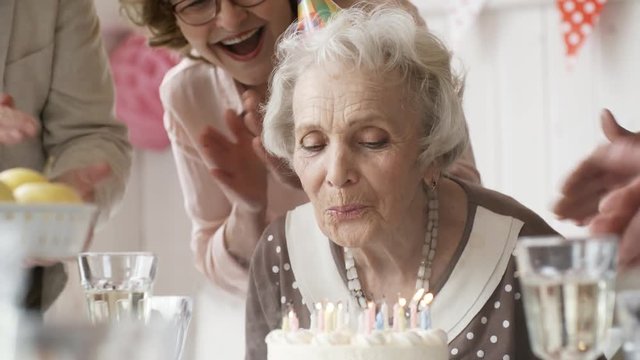 Beautiful senior woman  in party hat blowing off birthday candles on cake while joyous friends clapping hands and saying best wishes