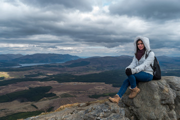 Naklejka premium Woman with fluffy white jacket and hiking shoes posing on a mountain with mountains and lakes in the backgroud, on a cloudy day at Nevis Range, Scottish Highlands