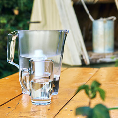 Water purification at countryside using water filter.
Water filter jug and a transparent cup of water with a village well on the background