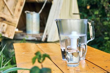 Water filter jug and a transparent cup of water standing on the wooden table with a open village well with a metallic bucket on the background outdoors
