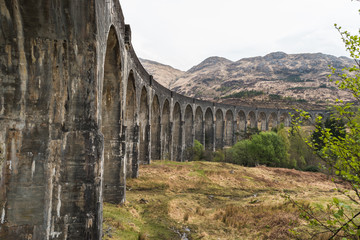Old train railway with arched bridge surrounded by trees with hills in view, Glennfinnan Viaduct, Scottish Highlands.